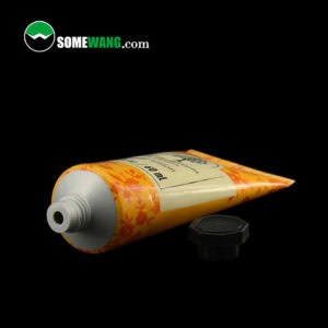 Một ống nhỏ bằng nhựa nhôm có đường kính 35mm chứa kem dưỡng da tay/kem dưỡng thể có nhãn hình chiếc lá màu cam và nắp lật màu đen, hiển thị trên nền tối.