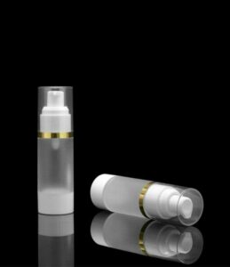 Two white 15/30/50/100ml Superior Quality AS Material Airless Pump Bottles with gold bands, one standing upright and the other lying down, on a reflective black surface.