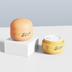 Two orange 30g cream jars, one upright on a white cube and one open, with "SW" label in a minimalist setting.