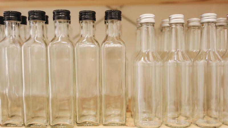 A row of clear glass bottles with black screw caps, arranged on a shelf. The bottles are uniform in size and shape, with a narrow neck and wider base, typical of beverage bottles. The focus is on the bottles in the foreground, with the background bottles becoming progressively blurred, creating depth.
