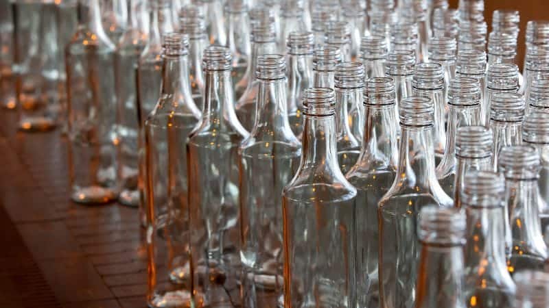 The image shows a large number of clear glass bottles arranged closely together on a wooden surface. These empty bottles have a uniform shape with a narrow neck and wider base, typical of beverage bottles. The focus is on the bottles in the foreground, with the background bottles becoming progressively blurred, creating depth.