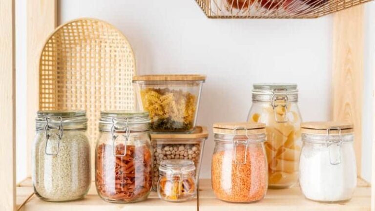 رف خشبي مُزود بعدة برطمانات زجاجية تحتوي على مكونات غذائية جافة. تتميز البرطمانات بأغطية معدنية محكمة الإغلاق تضمن نضارة الطعام. تشمل المحتويات الظاهرة الأرز والمعكرونة والبقوليات والتوابل، مما يُبرز استخدام البرطمانات الزجاجية في تنظيم المؤن وحفظ الطعام.