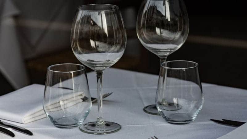 The glasses are set on a white linen tablecloth, which suggests a formal or fine dining setting. In the background, there is a folded white napkin with a set of cutlery on top of it, consisting of a fork and a knife, which are aligned parallel to each other. 