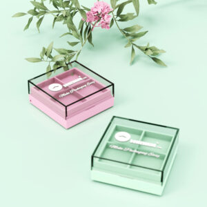 Deux palettes de fards à paupières de 4 couleurs (rose et vert avec couvercles transparents) sont posées près d'une fleur rose sur une surface vert clair.