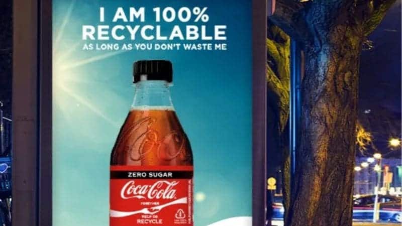 Cartelera que muestra un anuncio de Coca-Cola sin azúcar que enfatiza la reciclabilidad de la botella 100%, colocada junto a una calle de la ciudad por la noche.