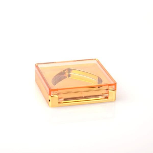 A small, square orange plastic blush case with mirror and a metallic triangular object sits on a plain white background.