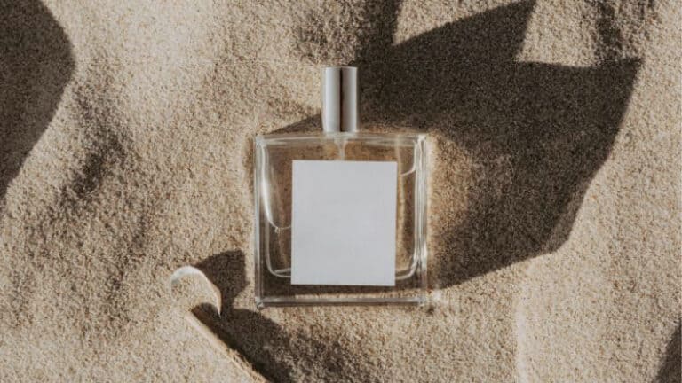 A square glass perfume bottle with a silver cap on coarse sand, next to a curved glass applicator, showcases elegant container design.