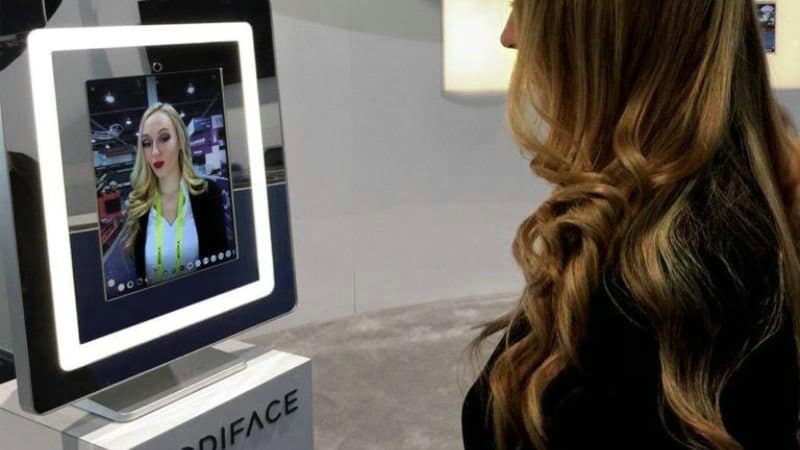 A woman with wavy hair stands before a digital screen showing her face, showcasing the latest beauty packaging trends.