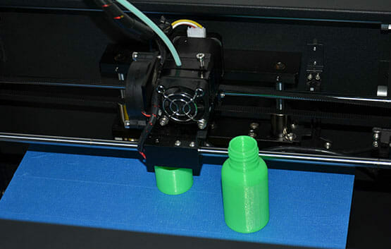 A 3D printer makes a green plastic bottle on a blue bed, with an identical finished bottle beside it.
